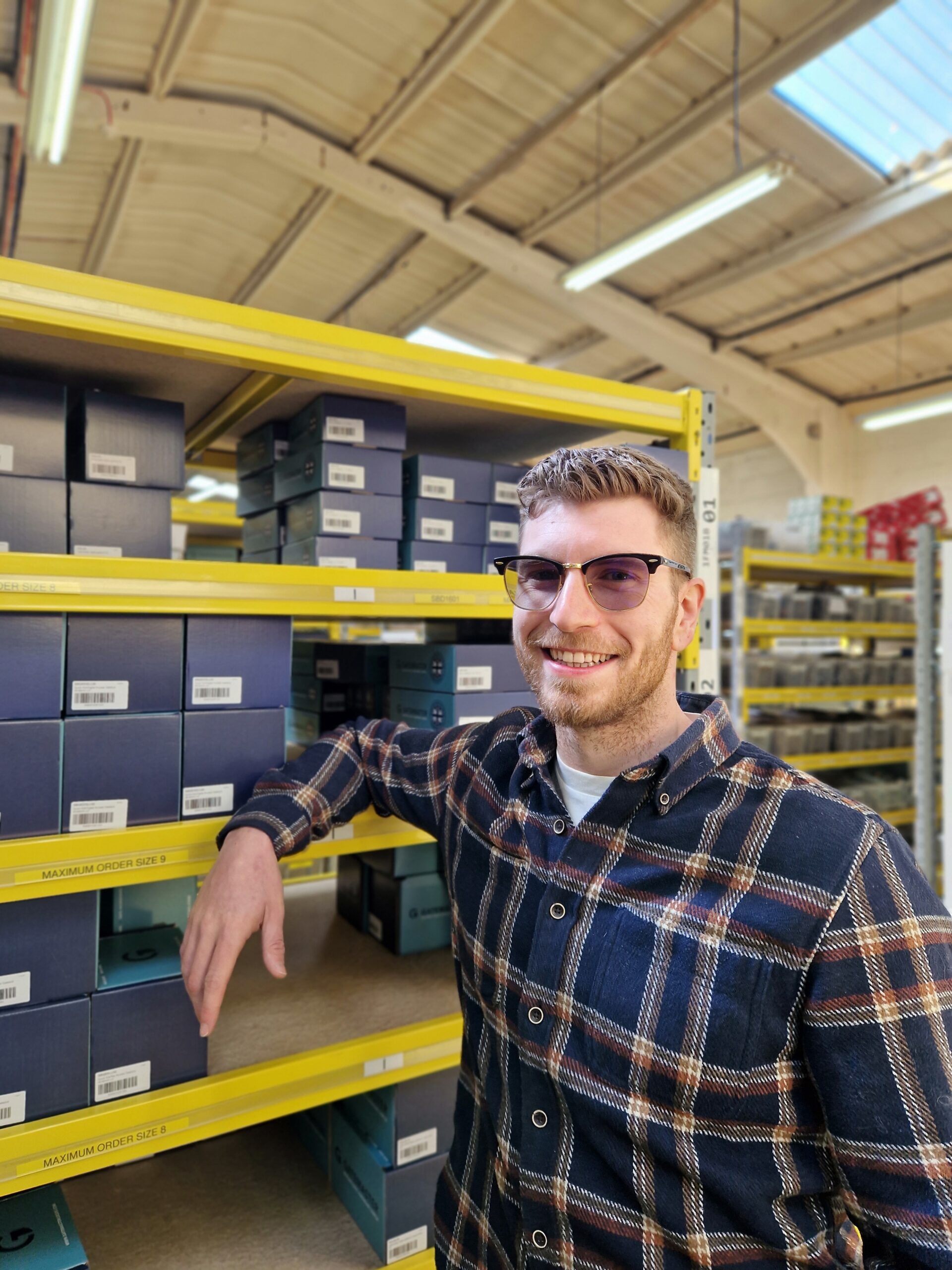 man in checked shirt with glasses smiling at camera in front of yellow racking.