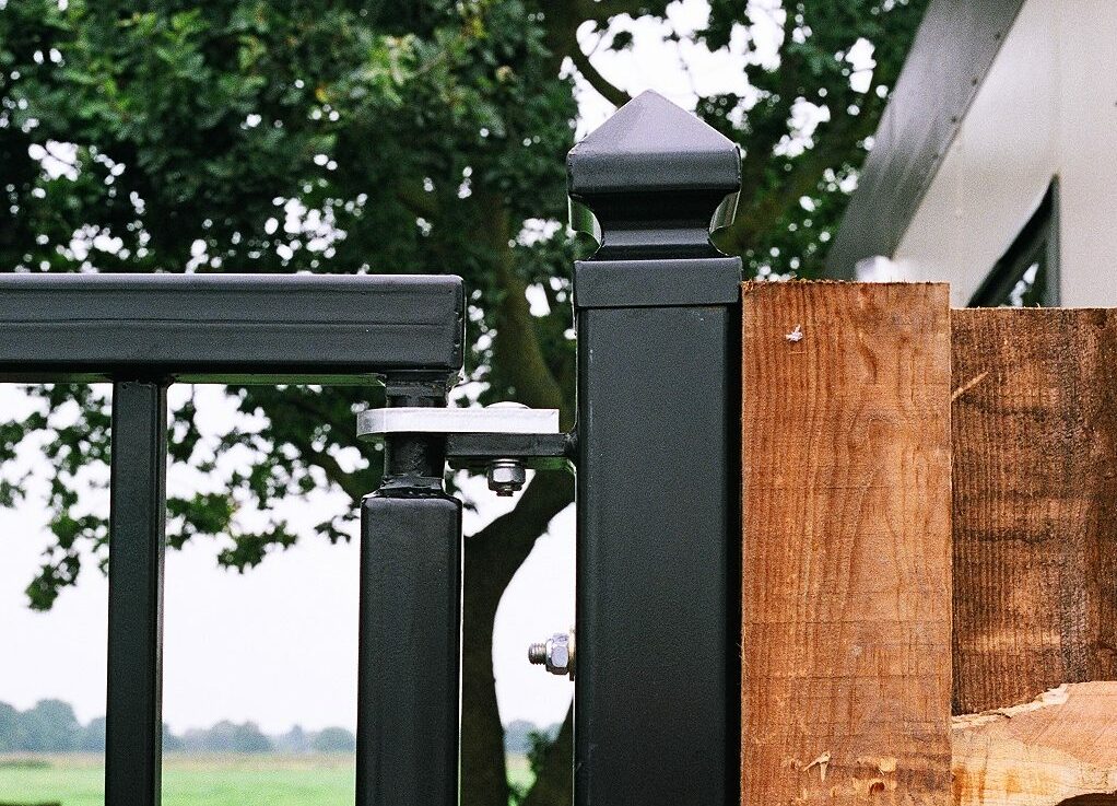Black metal gate post fixed to wooden fence. Top of metal gate has pivoting stainless steel gate hinge. In background: large green tree and fields