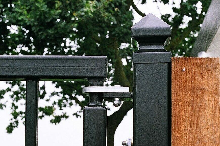 Black metal gate post fixed to wooden fence. Top of metal gate has pivoting stainless steel gate hinge. In background: large green tree and fields