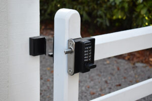 Image shows a black metal lock with a digital keypad fixed to a white wooden gate. In the background, there is a green hedge and a gravel path.