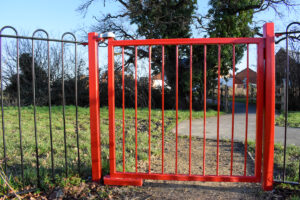 red pedestrian gate in park with bow top railings. Ensure safe gates for children with adjustable gate closer.