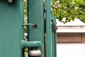 Close up of open green metal gate leaf with gate safety cable gate tether installed. Small braided steel wire going from gate post on left to gate leaf on right