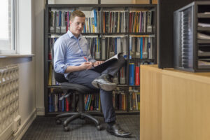 Man sitting in front of filled bookshelf. He has a pen and catalogue in his hands and one legs over the other