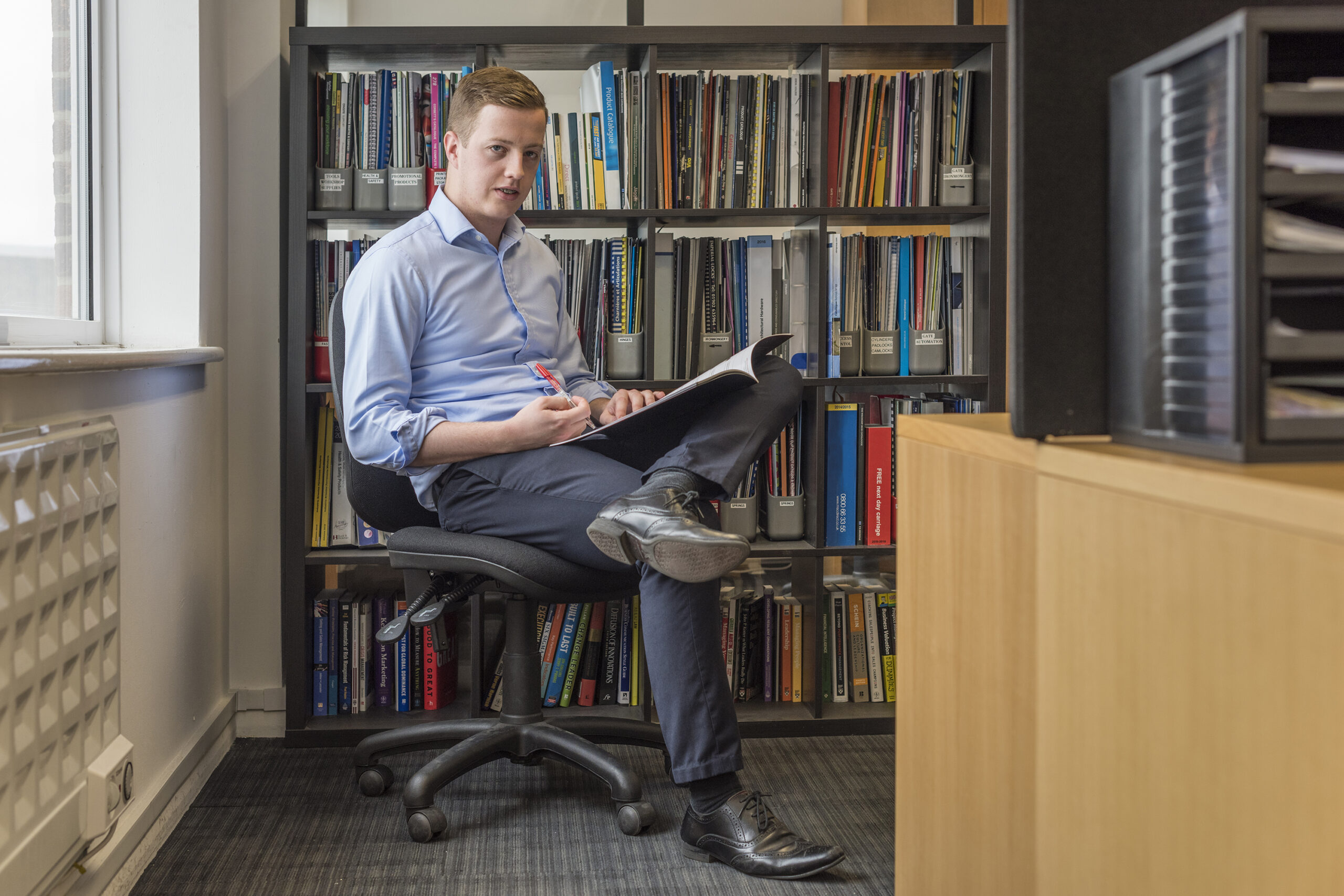 Man sitting in front of filled bookshelf. He has a pen and catalogue in his hands and one legs over the other