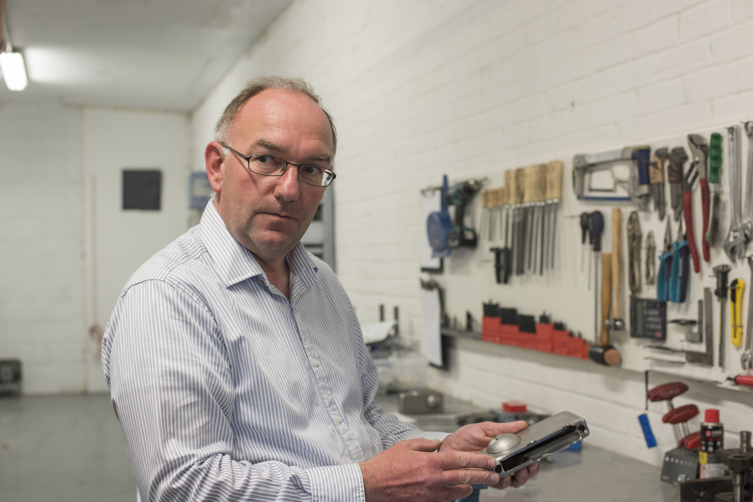 Portrait of man standing in front of work bench with tools. He is holding a metal lock case in his hand and looking to the left of camera