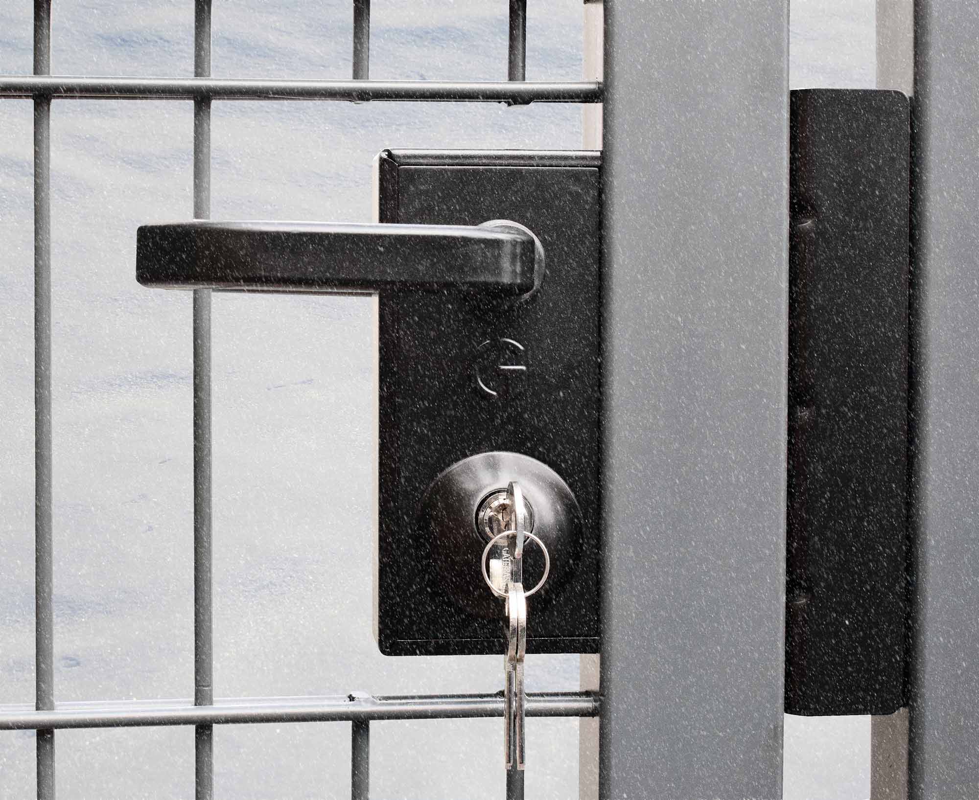 Black latch deadlock installed in metal gate. Small snowflakes in front and white snow banks behind the gate