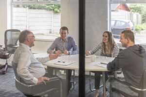 Three men and one woman sitting around a white table talking. Notepads, glasses and pens on the table