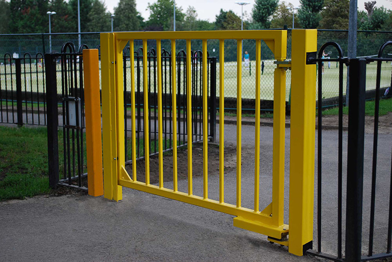 Yellow pedestrian gate with bowtop fencing on the sides. Installed in front of pathway and tennis courts in background